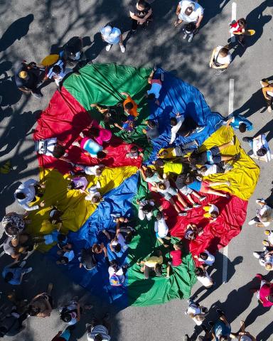 IMagem de drone. Do alto se vê crianças e adultos estendidos e sentados sobre um grande tecido colorido estendido na avenida. #paratodosverem 