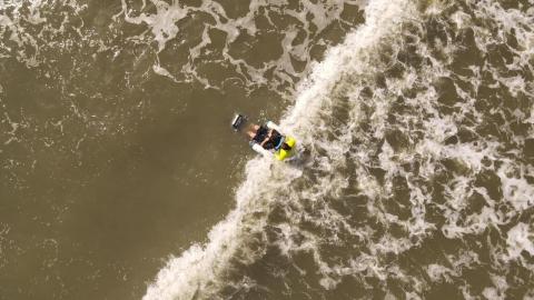 Imagem aérea por drone de pessoa tomando banho de mar em cadeira específica com apoio de condutor. #paratodosverem