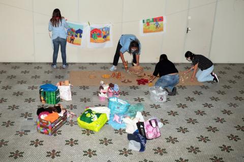 mulheres estão em salão distribuindo brinquedos no chão e afixando cartazes em parede. #paratodosverem