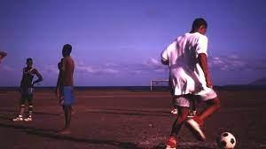 jovens em campo de areia jogam futebol #paratodosverem