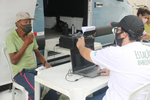 Dois homens sentados frente a frente em uma mesa. Um deles, diante de um notebook, fotografa o rosto do outro. #paratodosverem