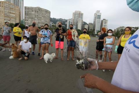 Em primeiro plano uma pessoa segura uma garrafa pet com resíduos sólidos dentro. Caes com tutores posam para foto ao fundo. #paratodosverem