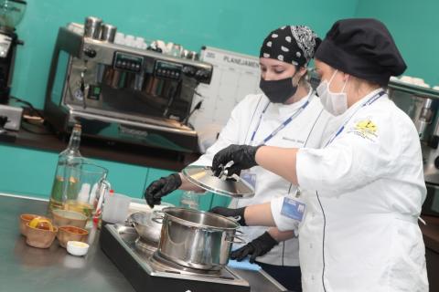 mulheres atrás de mesa preparam prato em panela. #paratodosverem