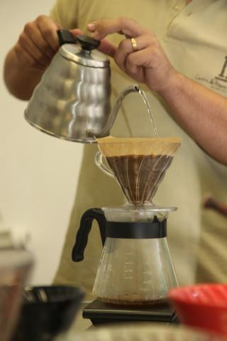 close de café sendo coado. Mãos seguram chaleira que despeja café no filtro. #paratodosverem