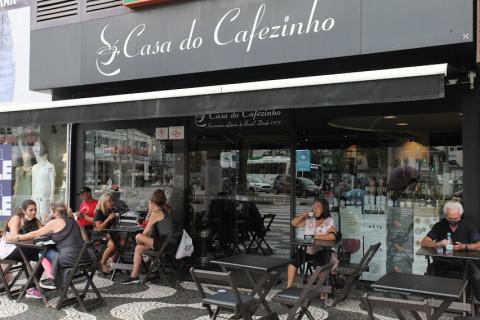Fachada da casa do cafezinho, com mesas e cadeiras na calçada onde algumas pessoas tomam café. #paratodosverem