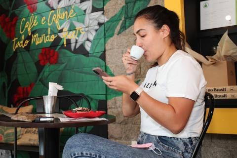 mulher toma cafezinho sentada a uma mesa. Uma das mmãos segura a xícara e a outra, o celular, para onde ela foca o olhar. #paratodosverem