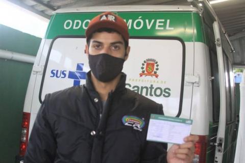 O caminhoneiro Caique Rafael exibe o comprovante de imunização. Atrás dele há uma ambulância estacionada. #para todosverem