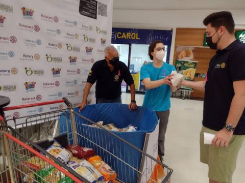 Pessoa entrega alimento ao lado de caixa de arrecadação e carrinho de supermercado cheio #paratodosverem