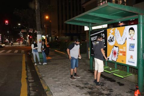 Duas pessoas afixam cartaz em ponto de ônibus a noite #paratodosverem