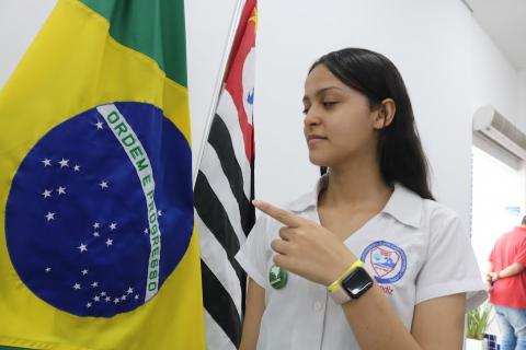 aprendiz uniformizada está apontando para a bandeira do Brasil. Atrás, a bandeira do estado de São Paulo 