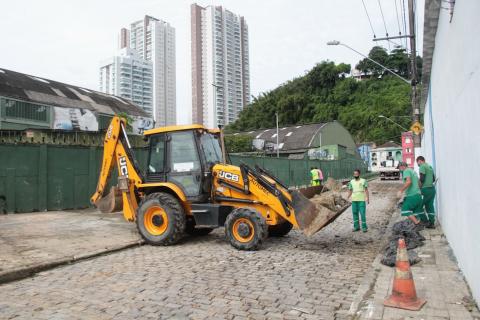 Retroescavadeira em primeiro plano na rua, com homens trabalhando. #Paratodosverem