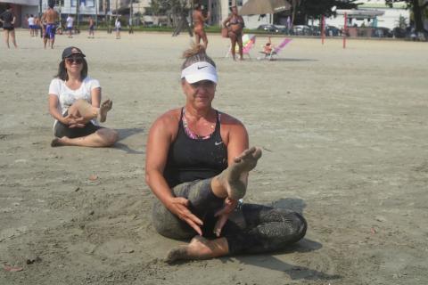 mulheres fazem exercício de aquecimento sentadas na areia da praia. #paratodosverem