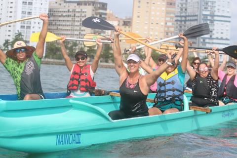 dentro da canoa e ainda na água, mulheres comemoram passeio com remos erguidos para o alto. #paratodosverem