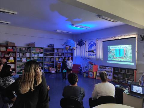 biblioteca com pessoas sentadas, em primeiro plano, e vídeo sendo exibido em projeção numa tela junto à parede. #paratodosverem