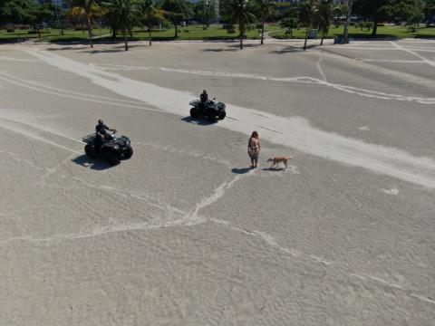 Guardas municipais em quadriciclos se aproximam de mulher que está na faixa de areia acompanhada de um cachorro. #Paratodosverem 