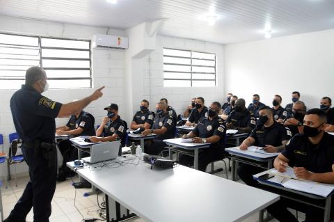 orientador atrás de mesa. Guardas municipais sentados olham para ele. #paratodosverem