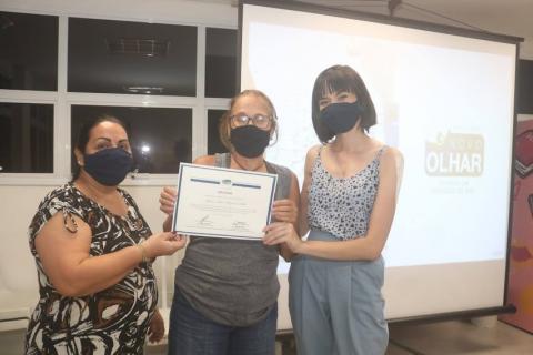 três mulheres posam para foto. A do meio segura um certificado. #paratodosverem