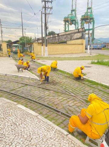 homens uniformizados estão agachados raspando grama entre os paralelepípedos no solo. Ao fundo, guindastes do porto. #paratodosverem