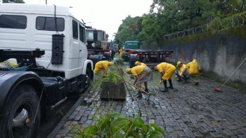 homens capinam calçada de paralelepípedos. Há caminhões estacionadas na guia à esquerda. #paratodosverem