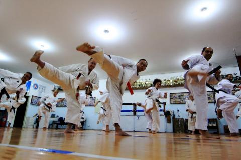 dojo de caratê com alunos dando golpe com as pernas. Os pés esquerdos estão no chão, e as pernas direitas no ar, em chute. Todos estão enfileirados. #paratodosverem