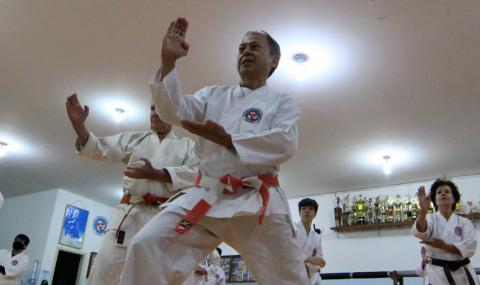 mestre Mitsu Shinzato, fazendo movimento de luta, com mãos em posição de defesa à frente do tronco. #paratodosverem