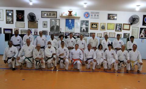 turma de alunos de caratê posa para foto. Uma parte está em pé ao fundo e outra, estão com as pernas flexionadas no chão. A parede tem inúmeros quadros e fotos.#paratodosverem