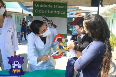Dentista mostra brinquedo com dentes para uma criança que está no colo da mãe. #paratodosverem