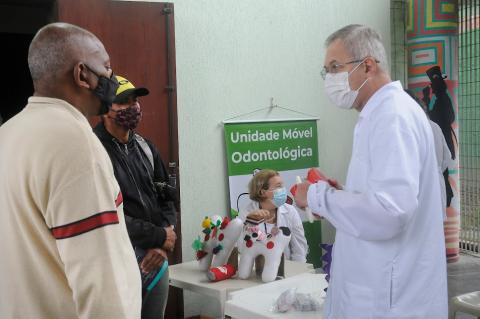 Homem usando avental branco conversa com outros dois homens no serviço de orientação odontológica. #paratodosverem