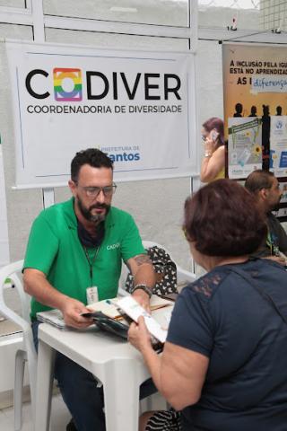 mulher está sentada, de costas, sendo atendida por um homem. Uma mesa separa os dois. Atrás dele há um banner onde se lê Codiver, da coordenadoria da diversidade. #paratodosverem