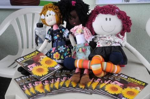 Três bonecas de pano sobre folhetos onde se lê: Ser criança. #paratodosverem