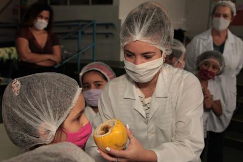 mulher segura maracujá e oferece para criança que está de máscara cheirar. Há outras crianças no ambiente. #paratodosverem