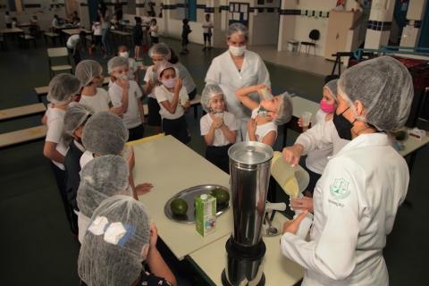 crianças estão reunidas em torno de mesa. Uma delas, ao fundo, bebe conteúdo de copo até o fim. Há um liquidificador na mesa. #paratodosverem