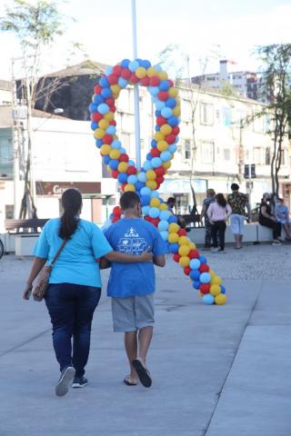 Mulher e jovem estão de costas caminhando em praça na direção do símbolo do autismo feito com bexigas coloridas. #paratodosverem