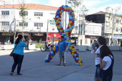 Pessoas circulam em praça. A esquerda, uma mulher fotografa que está à frente do símbolo do autismo feito com bexigas coloridas.#paratodosverem