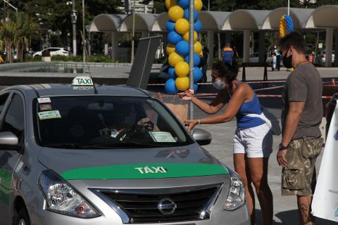 motorista de taxi e mulher do lado de fora do veículo seguram pacote com doações. #paratodosverem