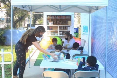 Crianças sentadas na mesa na frente da carreta lendo livros. #paratodosverem