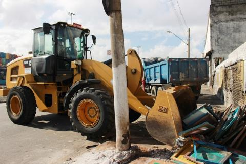 trator junta e recolhe inúmeros materiais junto a um muro. Há um caminhão para o despejo do material ao fundo. #paratodosverem