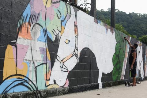 Muro grafitado, com desenho em primeiro plano de menina em cadeira de rodas, com os olhos fechados e a cabeça recostada sobre a cabeça do cavalo. #Paratodosverem