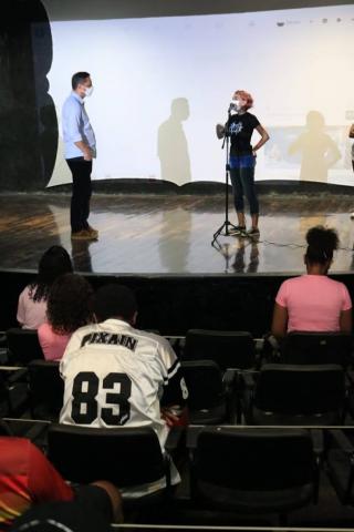 JOvem fala ao microfone no palco ao lado do prefeito. #paratodosverem