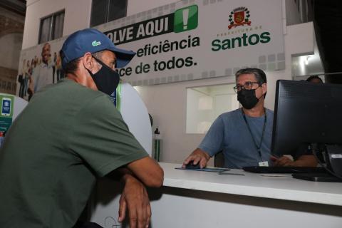 homem está sentado à mesa. Ele está de lado para a fota e diante de um balcão. Do outro lado, um homem à frente de um monitor de computador o atende. #paratodosverem