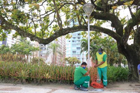 Homens instalam cerca plástica diante de jardim e poste de iluminação. #Paratodosverem