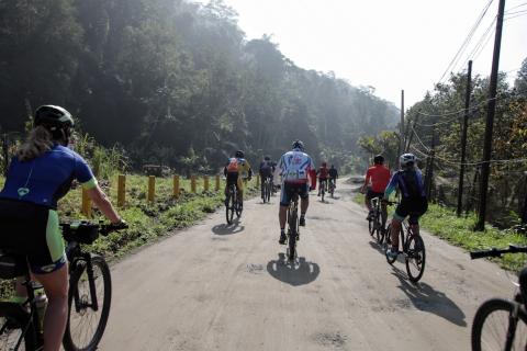 ciclistas pedalam em via asfaltada ladeada por ampla vegetação. #paratodosverem