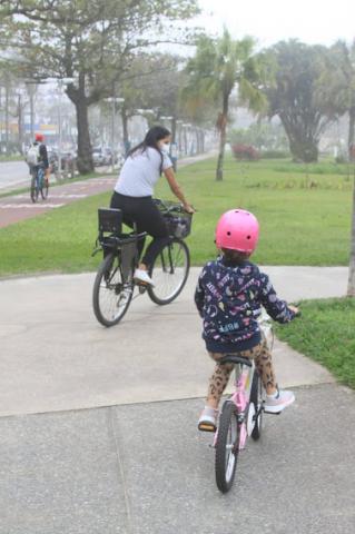 Mãe e filha pequena, de capacete, andando de bike no jardim da orla. #paratodosverem
