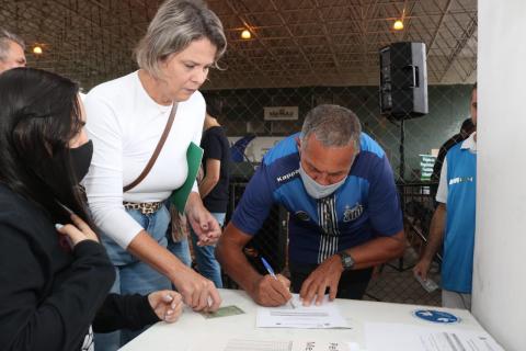 homem assina papel sobre uma mesa. Ao lado dele estão duas mulheres. #paratodosverem