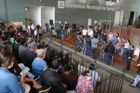 arquibancada de ginásio cheia de pessoas sentadas. Ao fundo, na quadra, movimentação de várias pessoas. Na parede da quadra se lê Prefeitura de Santos. #paratodosverem