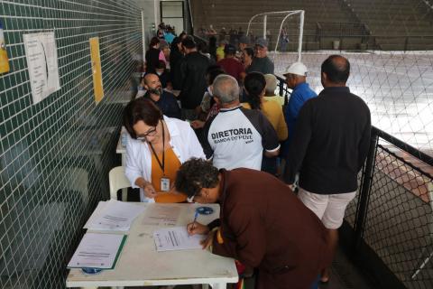 Mesa em primeiro plano com uma pessoa assinando documento. Há outras pessoas em torno. O ambiente é um corredor e háuma quadra esportiva ao lado. #paratodosverem