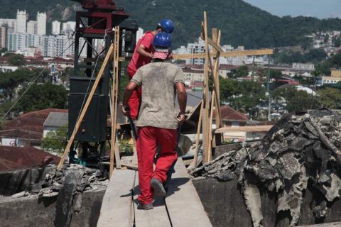 Homens trabalhando na cobertura. Dois deles passam sobre uma passarela de madeira. #Paratodosverem