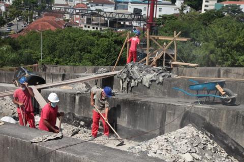 Homens trabalham na cobertura. Há estruturas de madeira aparente. Ao fundo se vê a vegetação. #Paratodosverem