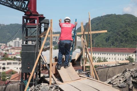 Homens trabalhando na cobertura. Um deles está parado com um carrinho de mão. Estrutura de madeira está aparente. #Paratodosverem