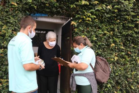 Dois agentes estão na porta de uma casa conversando com uma mulher. O muro é recoberto por plantas. #paratodosverem